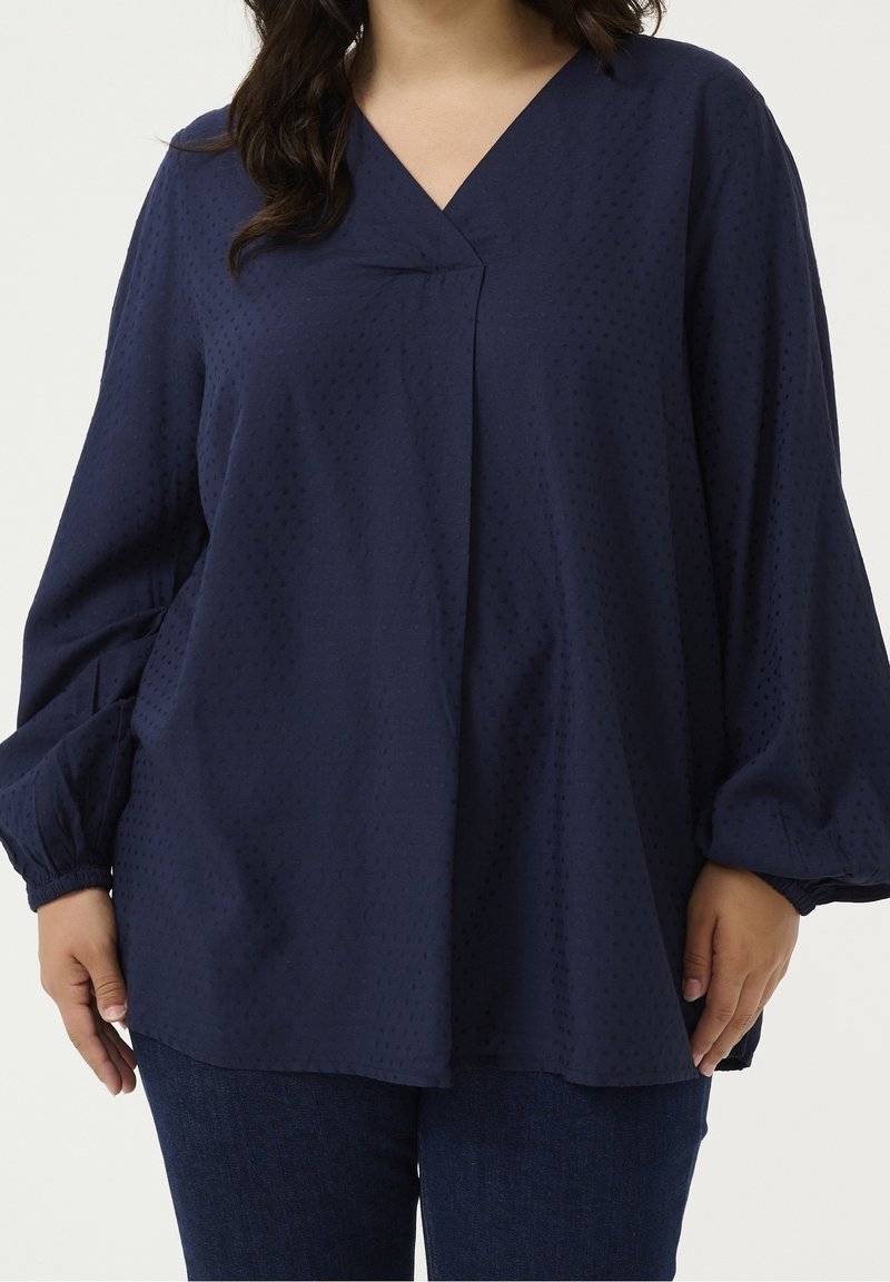 Femme portant un chemisier ample à manches longues bleu marine avec une texture subtilement poivrée et un col en V, associé à un jean bleu foncé.