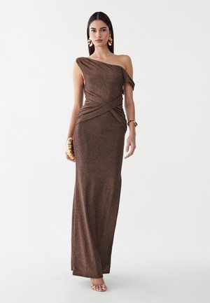 Vrouw draagt een bruine metallic avondjurk tot op de vloer met één schouder, houdt een gouden clutch vast, met gouden oorbellen en armband, staand tegen een witte achtergrond.