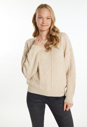Beige gestrickter Pullover mit Rauten- und Kabelmustern, langärmelig und mit Rippenabschluss. Kombiniert mit dunklen Jeans.