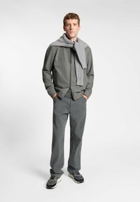 Giacca grigia con zip abbinata a pantaloni grigi e sneakers grigie. Sciarpa lavorata grigia con un motivo testurizzato. Sfondo piatto.