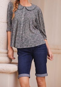 Zielona kwiecista bluzka z okrągłym kołnierzem, guzikami i plisowanymi detalami, zestawiona z ciemnoniebieskimi szortami jeansowymi podwiniętymi na końcach.