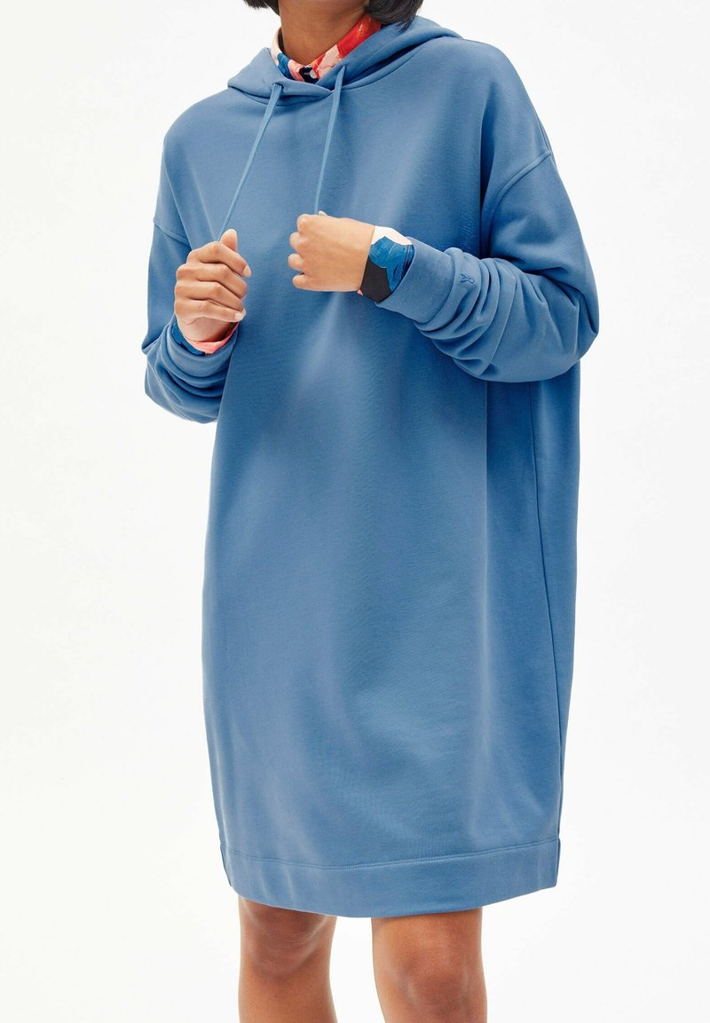 Personne portant une robe sweat à capuche bleue ample avec une manche de chemise colorée visible au poignet sur un fond blanc uni.