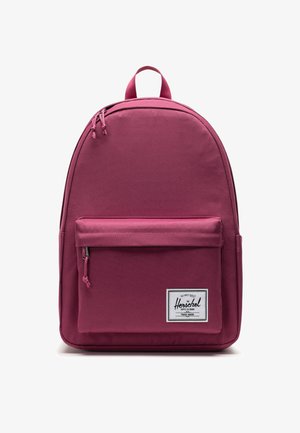 Sac à dos bordeaux avec une finition textile lisse, une forme arrondie, une poignée supérieure, une poche frontale et un patch logo blanc sur le bas.