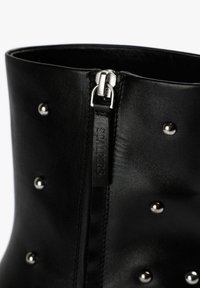 Bota de cuero negro para tobillo con tachuelas plateadas y una cremallera lateral, con una textura suave y una parte superior redondeada sin tacón.