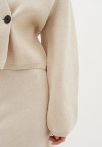 Cardigan bej tricotat, cu închidere prin buton, tivure ribbat, și manșete lungi, ușor bufante. Textura pare moale și confortabilă.
