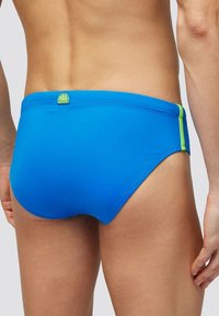 Bañador tipo slip azul hecho de material suave con costuras verdes en contraste. Presenta una etiqueta con el logo situada en la parte posterior.