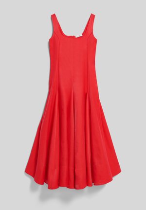 Kvinde iført rød midikjole uden ærmer med firkantet halsudskæring og sorte hænderør med nitter, stående foran en ensfarvet grå baggrund.