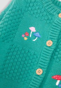 Cardigan en tricot turquoise avec un motif texturé, des boutons en bois et des champignons brodés colorés avec des fleurs.