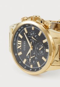 Guldfarvet kronografur med sort urskive, fremtrædende time markører, sub-ur og tachymeter. Har poleret metalrem og krone.