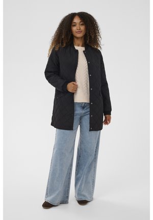 Melnā quiltota jaka ar pogām, ribotām piedurknēm un brīvu piegriezumu, valkāta virs bežīgas adītas džemperis un gaišzilas plašas bikses.