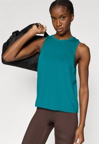 Τιρκουάζ αμάνικη αθλητική μπλούζα από λείο ύφασμα, με στρογγυλή λαιμόκοψη και ελαφρώς φαρδιά εφαρμογή, συνδυασμένη με καφέ στενά κολάν.