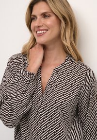 Camicia a righe marrone e bianca con scollatura a V e maniche lunghe, caratterizzata da un design geometrico e una texture di tessuto morbido.