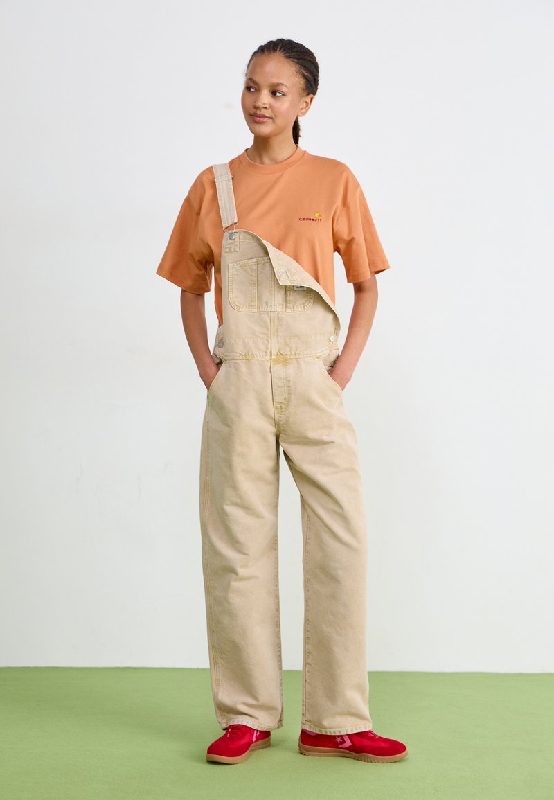 Beżowe ogrodniczki dżinsowe z przednią kieszenią, noszone na pomarańczowej koszuli z małym logo. Uzupełnione czerwonymi trampkami na zielonej podłodze.