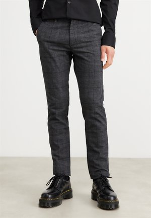Mann trägt schlanke graue karierte Hose und schwarze klobige Schnürschuhe, steht vor einfachem Hintergrund.