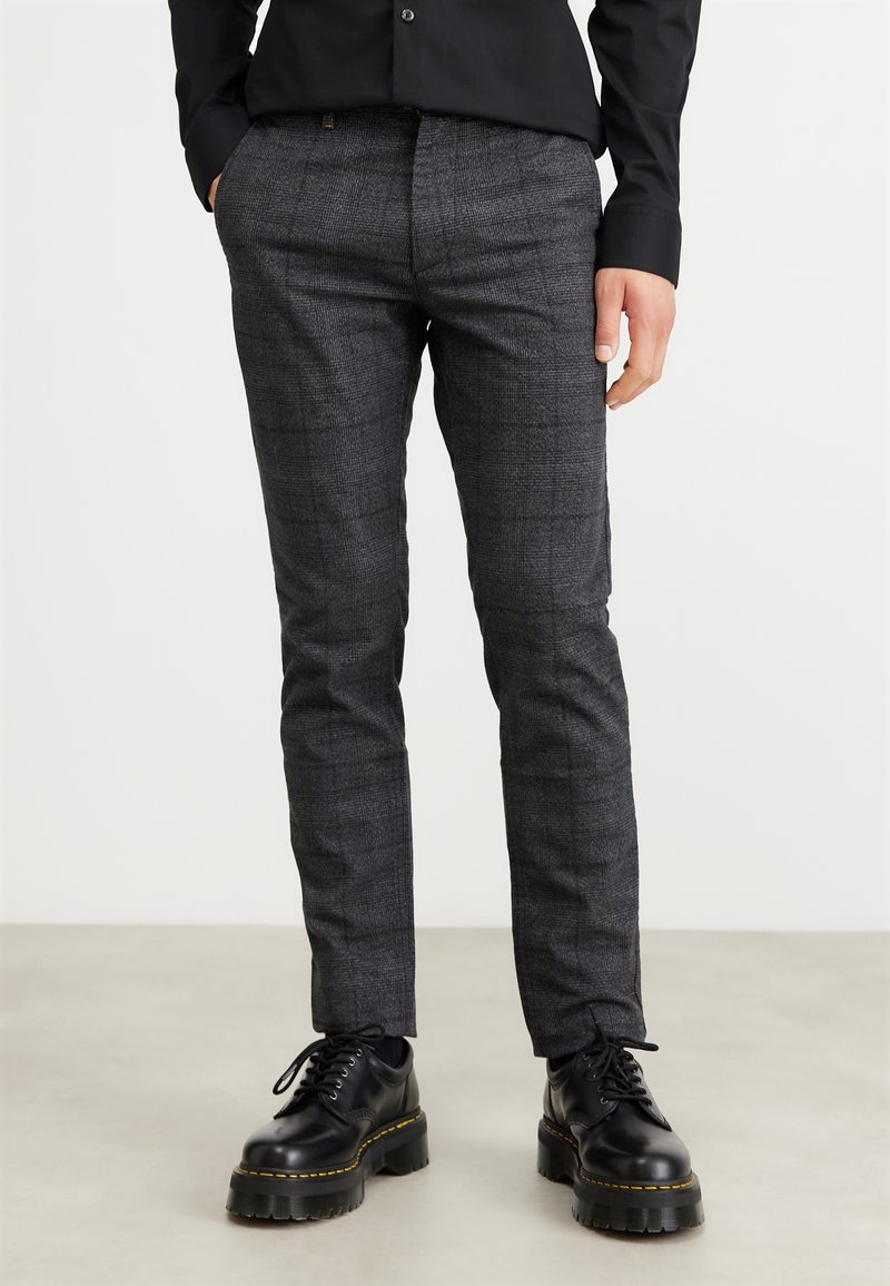 Homme portant un pantalon slim gris à carreaux et des chaussures noires épaisses à lacets, debout devant un fond uni.