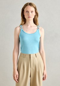 Light blue tank top with shiny texture and decorative blue hardware on straps, paired with beige high-waisted, pleated trousers.