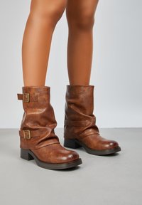 Bottines en cuir marron à design décontracté, dotées de deux sangles à boucle dorée et d'un talon épais. Texture lisse et orteils arrondis.