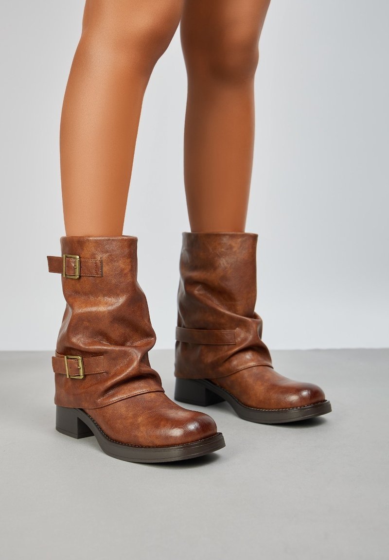Bottines en cuir marron à design décontracté, dotées de deux sangles à boucle dorée et d'un talon épais. Texture lisse et orteils arrondis.