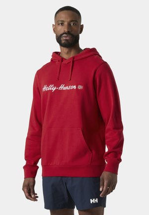 Man draagt een rode Helly-Hansen hoodie met logo en marineblauwe shorts met HH-logo tegen een effen achtergrond.