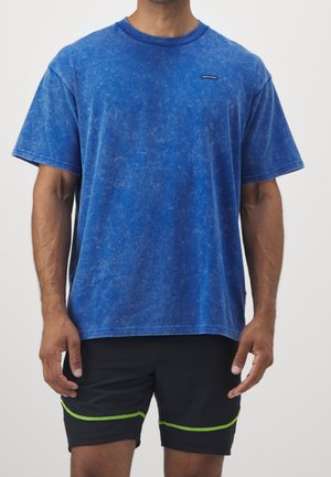 Homme portant un T-shirt bleu à manches courtes lavé et un short noir avec des coutures vertes, debout devant un fond uni.