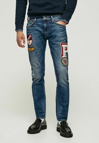 Jeans en denim bleu avec des patches comportant des logos et du texte. Zones décolorées et légères usures. Conçus avec une coupe droite et un style classique à cinq poches.