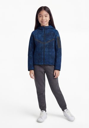 Jeune fille debout et souriante, portant une veste à motif bleue avec fermeture éclair, un pantalon gris foncé et des baskets blanches.
