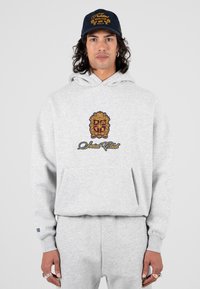 Felpa grigia con un grande emblema ricamato in rosso scuro e oro, tasca anteriore a marsupio, abbinata a pantaloni della tuta coordinati. Cappellino blu navy con logo.