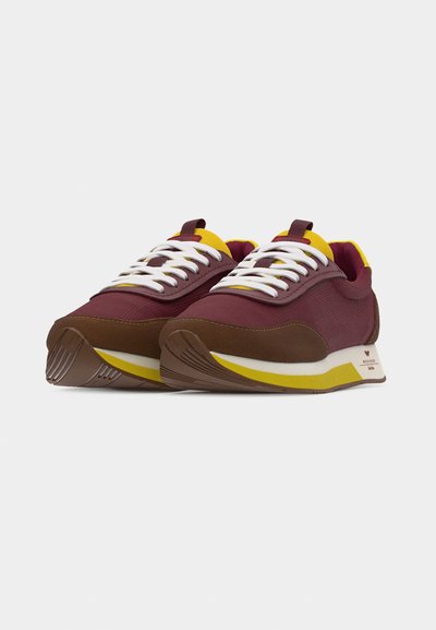 Paire de baskets bordeaux et marron avec lacets blancs, accents jaunes et semelles amortissantes, inclinées pour montrer l'avant et le côté extérieur.