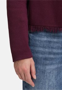 Burgunderfarbener Strickpullover mit Fransensaum und langen Ärmeln. Kombiniert mit blauer Denim, die einen strukturierten Stoff und eine lässige Passform zeigt.