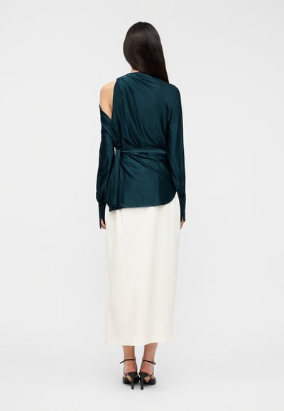 Γυναίκα με μακριά σκούρα μαλλιά φοράει σκούρο τιρκουάζ μπλούζα που δένει, με έναν ώμο ακάλυπτο, λευκή midi φούστα και μαύρα σανδάλια με τακούνι, κοιτάζει προς τα πίσω.