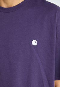 Fialové bavlnené tričko s bielym logom na ľavej strane hrude. Krátke rukávy, okrúhly výstrih, jemná textúra.