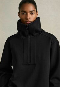 Felpa nera con colletto alto e zip frontale. Caratterizzata da una texture liscia e un design strutturato con dettagli di cucitura delicati.
