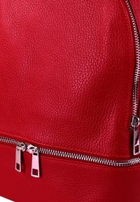 Sac à dos en cuir rouge avec une surface texturée, muni d'une fermeture éclair en métal argenté et de deux tirettes au bas pour une fermeture sécurisée.