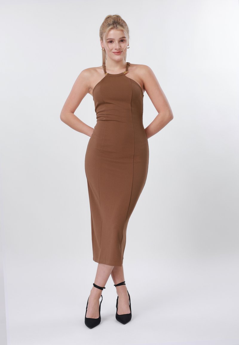 Γυναίκα που στέκεται με τα χέρια πίσω από την πλάτη, φορώντας ένα αμάνικο καφέ μίντι φόρεμα και μαύρες γόβες με λουράκι στον αστράγαλο, σε απλό λευκό φόντο.