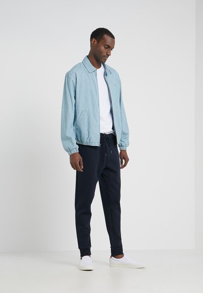 Veste en denim bleu clair avec col, fermeture éclair à l'avant et poignets élastiques, associée à un jogger bleu marine foncé et des chaussures blanches à enfiler.