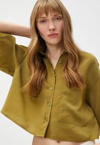 Olijfgroene, korte button-up blouse met korte mouwen, voorzak en subtiele glans. Heeft groene knopen en een ontspannen pasvorm.