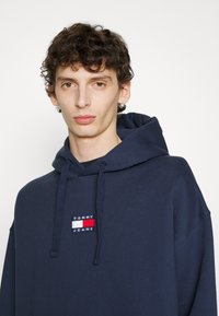 Felpa con cappuccio di colore blu navy, realizzata in cotone, con un logo ricamato "TOMMY JEANS" e strisce in rosso, bianco e blu.