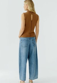 Bruine mouwloze top met een hoge hals en een keyhole-detail op de rug, gecombineerd met lichtblauwe brede jeans met achterzakken en een ontspannen pasvorm.