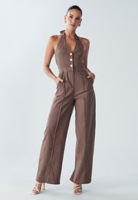Bruine linnen jumpsuit met halterhalslijn, vier knopen en zijzakken. Voorzien van wijde pijpen en minimalistisch ontwerp.