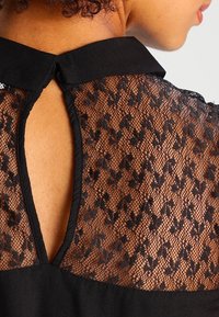 Blouse noire avec col et ouverture en forme de goutte dans le dos. Présente des détails en dentelle avec un motif floral. Le tissu texturé contraste avec le col lisse.