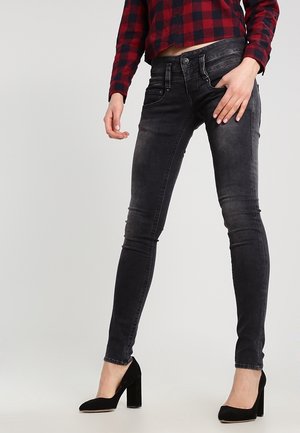 Osoba w wyblakłych czarnych obcisłych dżinsach, czarnych butach na słupku i krótkiej czerwono-czarnej koszuli w kratę, stojąca na jednolitym tle.