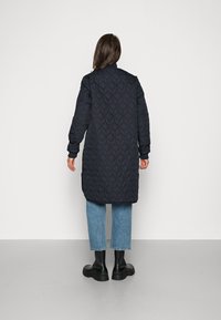 Cappotto lungo, blu scuro, trapuntato con un motivo a diamante, polsini a coste e colletto alto. Indossato sopra jeans azzurri chiari e stivali neri.