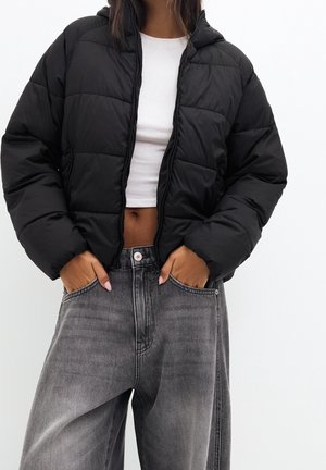 Femme portant une doudoune noire par-dessus un crop top blanc et un jean gris ample, les mains dans les poches du jean.