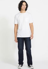 Biały bawełniany t-shirt z kieszonką na piersi, w połączeniu z ciemnoniebieskimi dżinsami i czarnymi trampkami. Model stoi z neutralnym wyrazem twarzy.