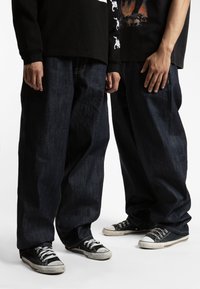 Jeans baggy in denim blu scuro con dettagli di cucitura visibili, abbinati a sneakers in tela nere con lacci bianchi e suole in gomma.