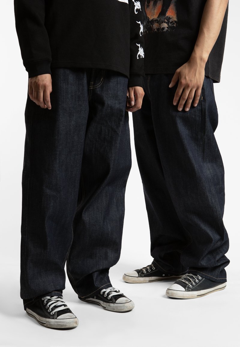 Jeans baggy in denim blu scuro con dettagli di cucitura visibili, abbinati a sneakers in tela nere con lacci bianchi e suole in gomma.