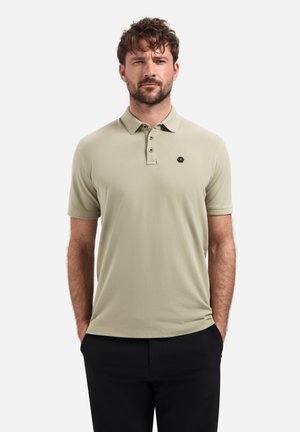 Mann mit kurzen braunen Haaren und Bart, der ein beigefarbenes kurzärmeliges Poloshirt und schwarze Hose trägt, mit den Händen in den Taschen stehend.