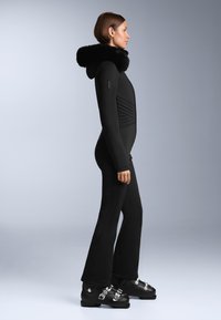 Czarny kombinezon narciarski z wysokim kołnierzem, posiadający futrzany kaptur, prążkowane akcenty i rozszerzane nogawki. W połączeniu z czarnymi butami narciarskimi i srebrnymi elementami.