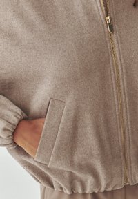Světle hnědá bomber bunda s měkkou strukturou, se zipem, elastickými manžetami a boční kapsou. Má uvolněný střih a minimalistický design.