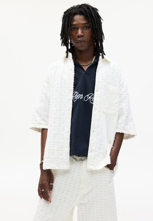 White textured shirt with short sleeves, paired with matching patterned pants. Underneath is a dark blue polo featuring white text.